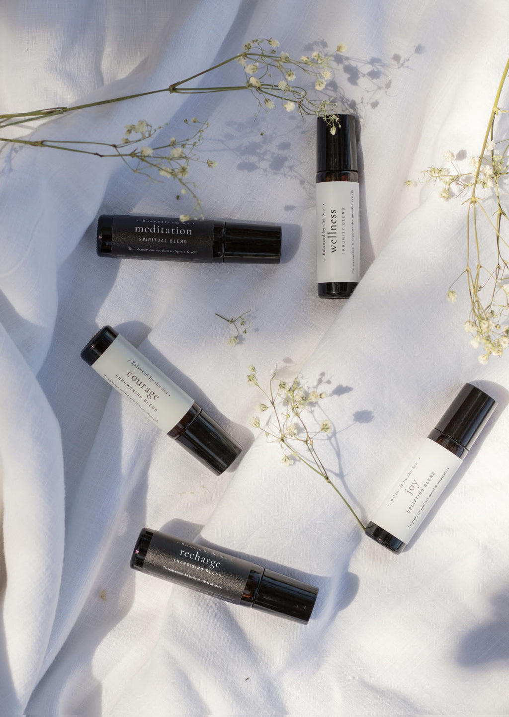 Five amber Balanced by the Sea essential oil roller bottles are displayed on a white surface with a white marble background.