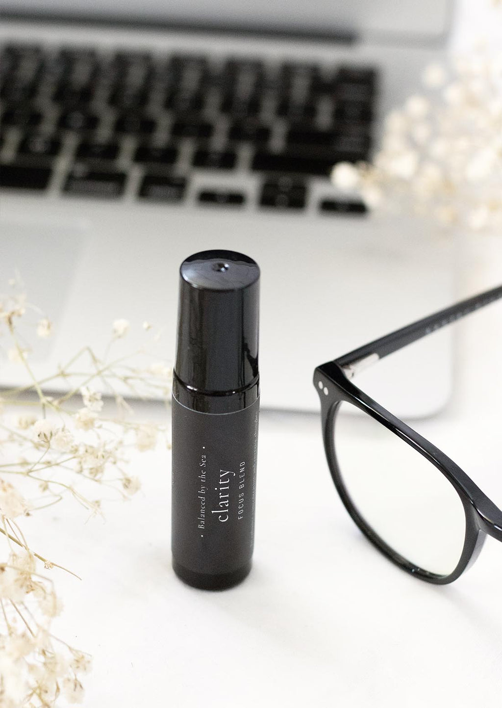 An amber essential oil roller bottle is displayed  on a white surface with a white marble background. It has a black cap and a black Balanced by the Sea label with the words "Clarity - Focus Blend" on it