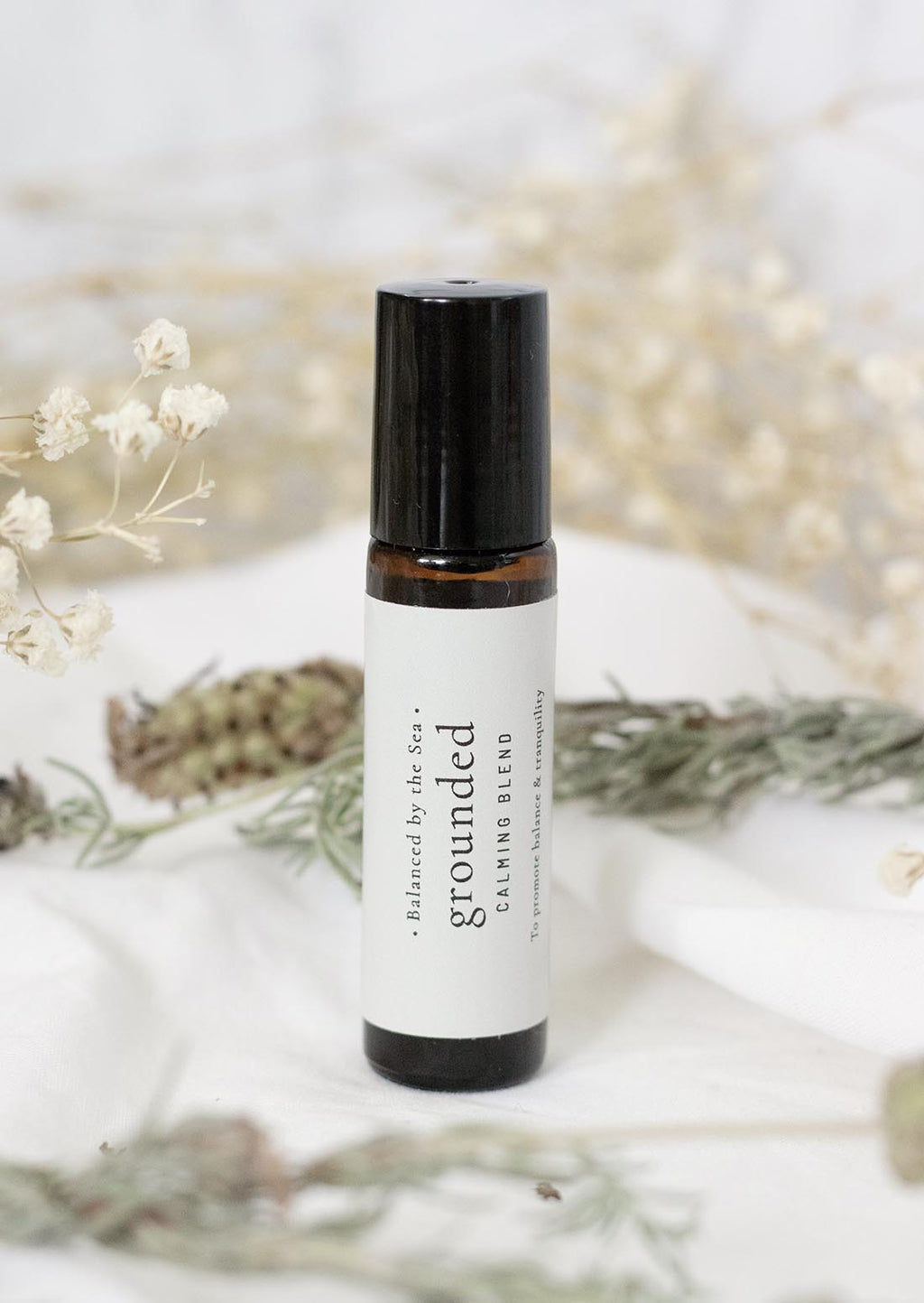 An amber essential oil roller bottle is displayed on a white surface with a white marble background. It has a black cap and a white Balanced by the Sea label with the words "Grounded - Calming Blend" on it