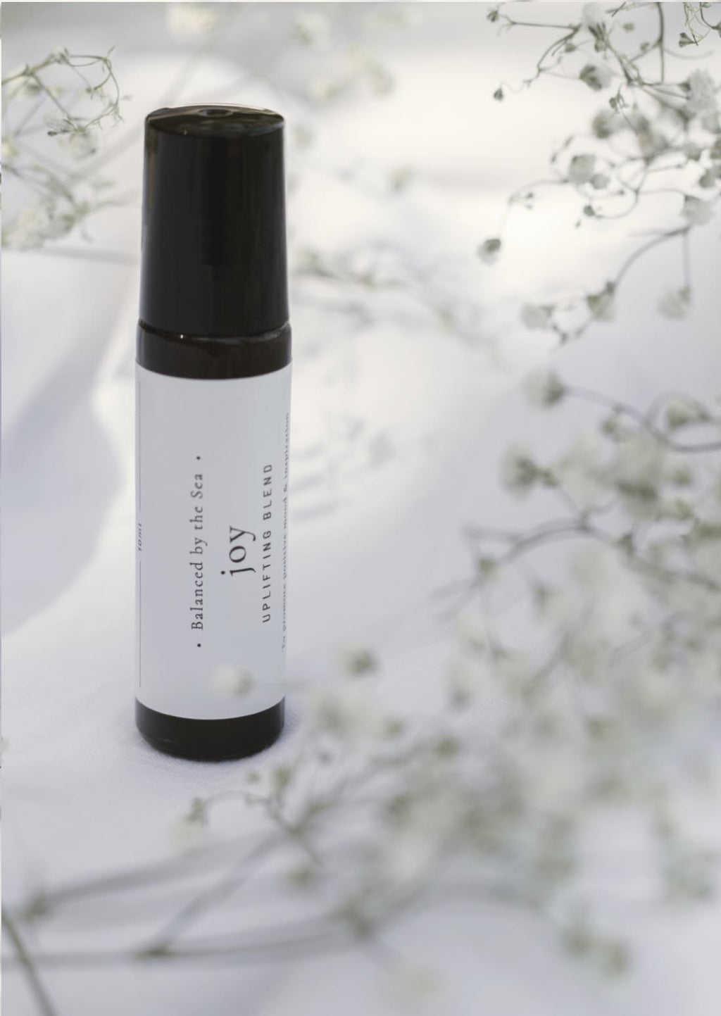 An amber essential oil roller bottle is displayed on a white surface with a white marble background. It has a black cap and a white Balanced by the Sea label with the words "Joy - Uplifting Blend" on it