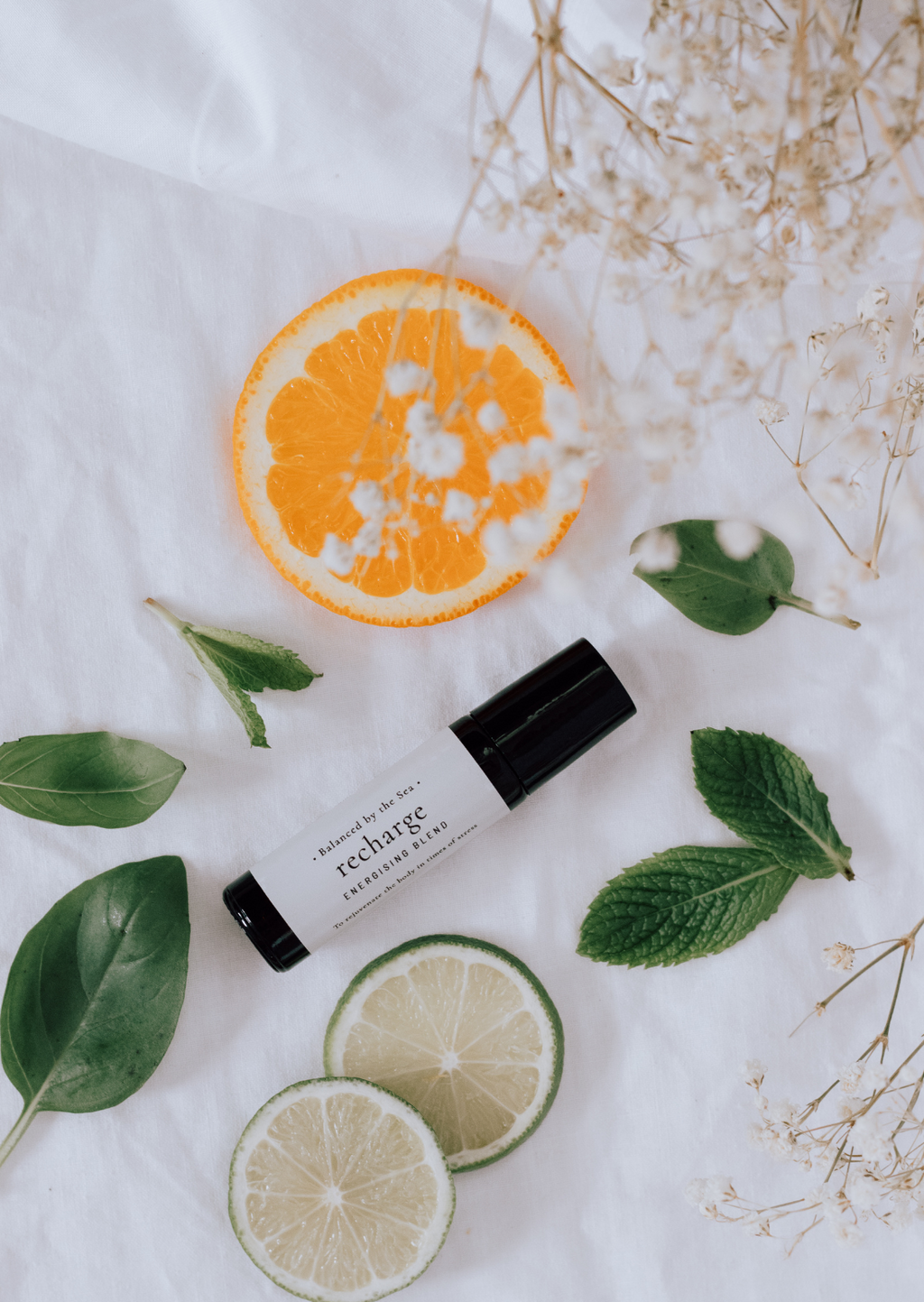 An amber essential oil roller bottle is displayed on a white surface with a white marble background. It has a black cap and a white Balanced by the Sea label with the words "Recharge - Energising Blend" on it