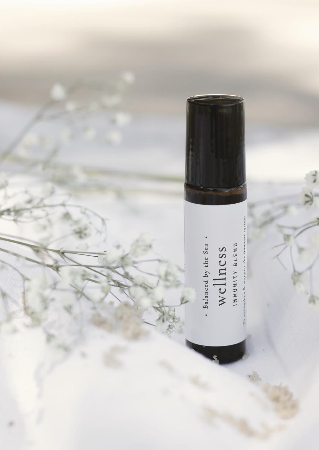 An amber essential oil roller bottle is displayed on a white surface with a white marble background. It has a black cap and a white Balanced by the Sea label with the words "Wellness - Immunity Blend" on it