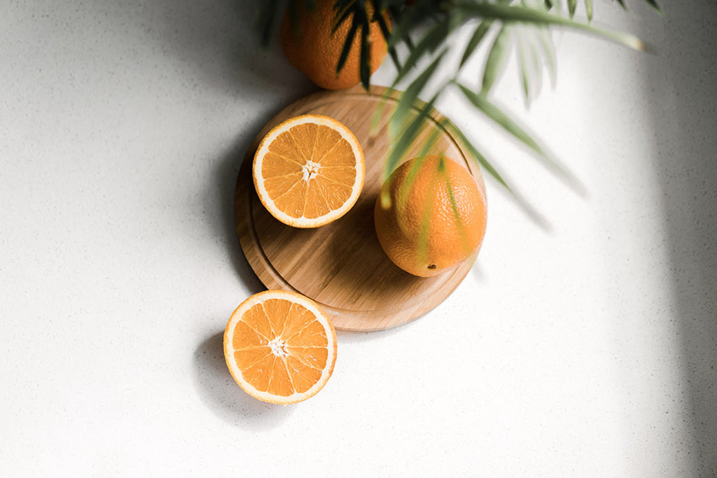 a whole orange and two orange halves are arranges on a wooden centre piece that has been place on a white tablecloth. There is a palm leaf hanging over them