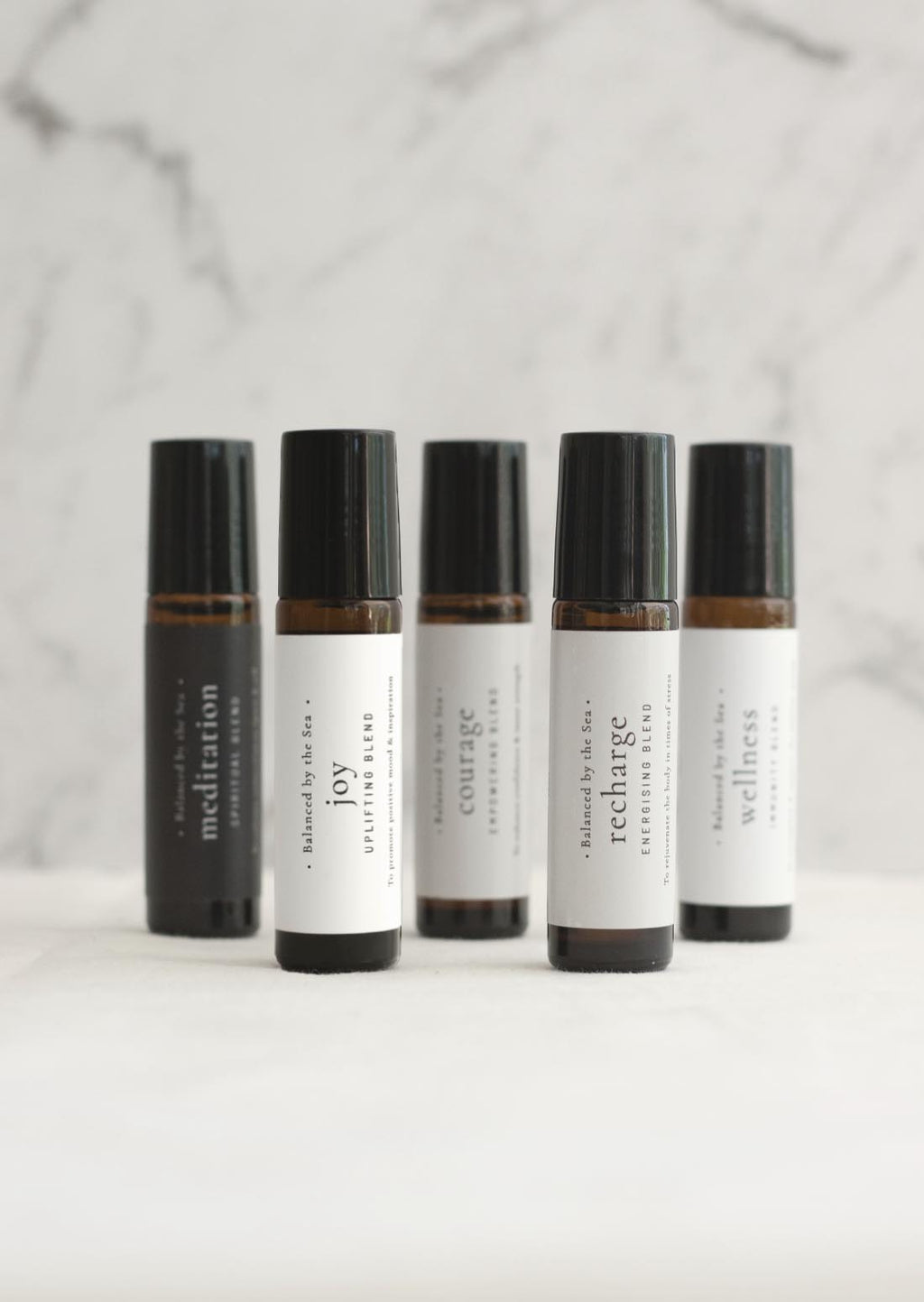 Five amber Balanced by the Sea essential oil roller bottles are displayed on a white surface with a white marble background.