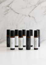 Nine amber Balanced by the Sea essential oil roller bottles are displayed on a white surface with a white marble background.