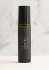 An amber essential oil roller bottle is displayed  on a white surface with a white marble background. It has a black cap and a black Balanced by the Sea label with the words "Clarity - Focus Blend" on it