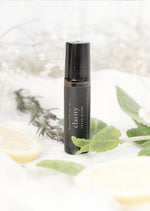 An amber essential oil roller bottle is standing on a white tablecloth, surrounded by slices of lemon, rosemary stems, mint leaves and fine white gypsum flowers. It has a black cap and a black Balanced by the Sea label with the words "Clarity - Focus Blend" on it