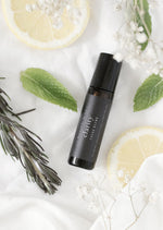 An amber essential oil roller bottle is lying on a white tablecloth, surrounded by slices of lemon, rosemary stems, mint leaves and fine white gypsum flowers. It has a black cap and a black Balanced by the Sea label with the words "Clarity - Focus Blend" on it