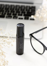 An amber essential oil roller bottle is standing on a white tablecloth, surrounded fine white gypsum flowers. It has a black cap and a black Balanced by the Sea label with the words "Clarity - Focus Blend" on it. In the background a laptop and a pair of black glasses can be seen.