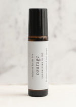 An amber essential oil roller bottle is displayed on a white surface with a white marble background. It has a black cap and a white Balanced by the Sea label with the words "Courage - Empowering Blend" on it