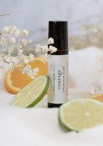 An amber essential oil roller bottle is standing on a white tablecloth, surrounded by slices of lime and orange and fine white gypsum flowers. It has a black cap and a white Balanced by the Sea label with the words "Courage - Empowering Blend" on it