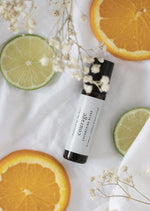 An amber essential oil roller bottle is lying on a white tablecloth, surrounded by slices of lime and orange and fine white gypsum flowers. It has a black cap and a white Balanced by the Sea label with the words "Courage - Empowering Blend" on it