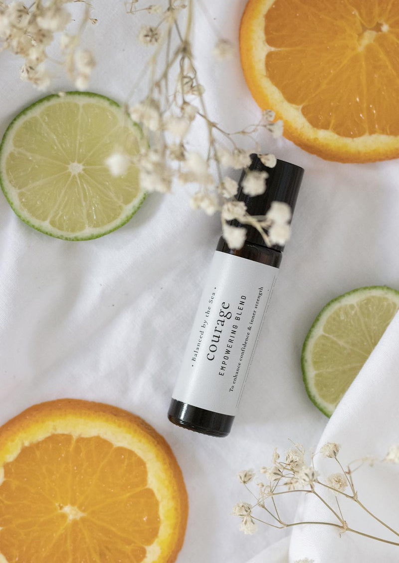 An amber essential oil roller bottle is lying on a white tablecloth, surrounded by slices of lime and orange and fine white gypsum flowers. It has a black cap and a white Balanced by the Sea label with the words "Courage - Empowering Blend" on it