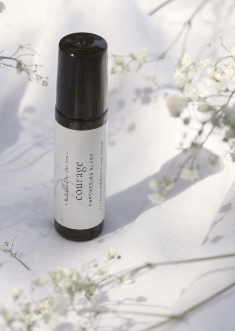 An amber essential oil roller bottle is standing on a white tablecloth, surrounded by fine white gypsum flowers. It has a black cap and a white Balanced by the Sea label with the words "Courage - Empowering Blend" on it