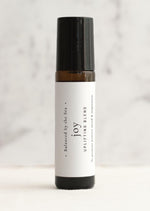 An amber essential oil roller bottle is displayed on a white surface with a white marble background. It has a black cap and a white Balanced by the Sea label with the words "Joy - Uplifting Blend" on it