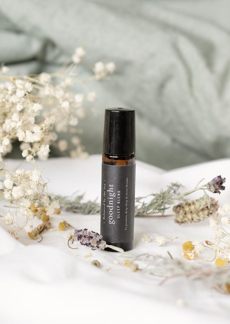 An amber essential oil roller bottle is standing on a white quilt, surrounded by stems of lavender, chamomile flowers and fine white gypsum flowers. It has a black cap and a black Balanced by the Sea label with the words "Goodnight - Sleep Blend" on it