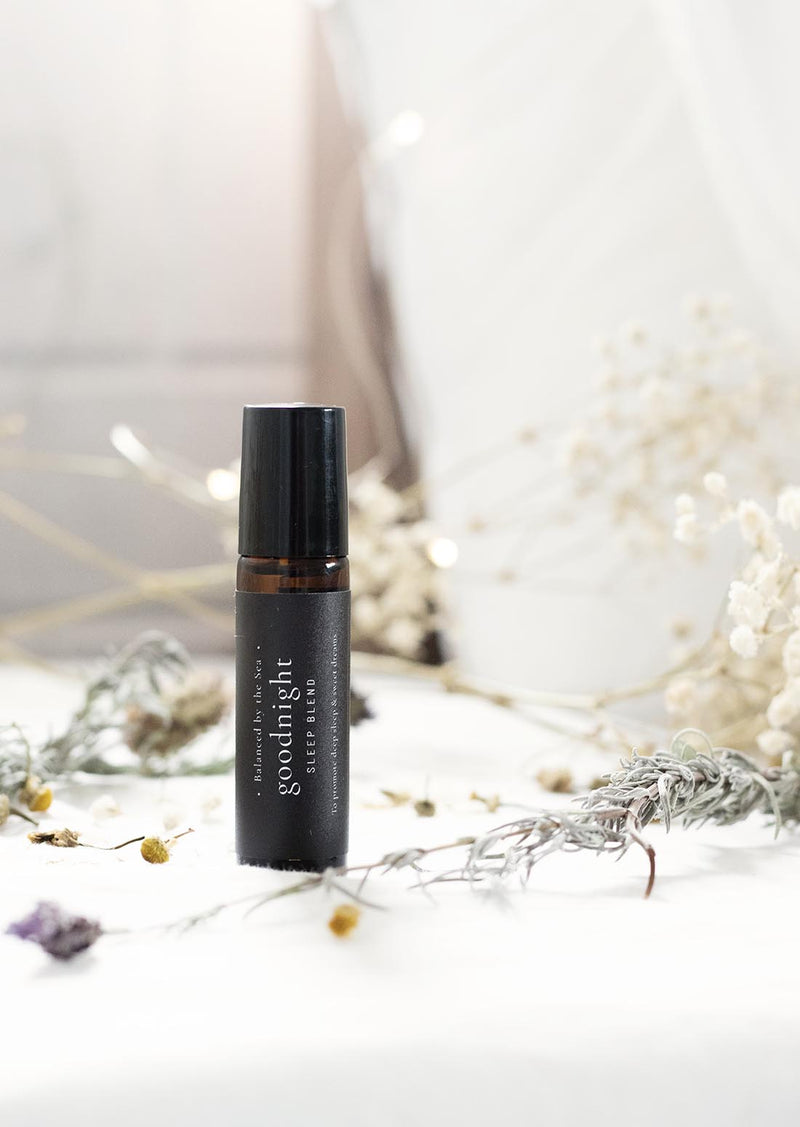 An amber essential oil roller bottle is standing on a white quilt, surrounded by stems of lavender, chamomile flowers and fine white gypsum flowers. It has a black cap and a black Balanced by the Sea label with the words "Goodnight - Sleep Blend" on it