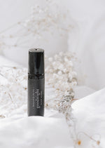 An amber essential oil roller bottle is standing on a white quilt, surrounded by stems of lavender, chamomile flowers and fine white gypsum flowers. It has a black cap and a black Balanced by the Sea label with the words "Goodnight - Sleep Blend" on it