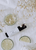 An amber essential oil roller bottle is lying on a white tablecloth, surrounded slices of lime and fine white gypsum flowers. It has a black cap and a white Balanced by the Sea label with the words "Joy - Uplifting Blend" on it