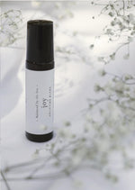 An amber essential oil roller bottle is standing on a white tablecloth, surrounded fine white gypsum flowers. It has a black cap and a white Balanced by the Sea label with the words "Joy - Uplifting Blend" on it