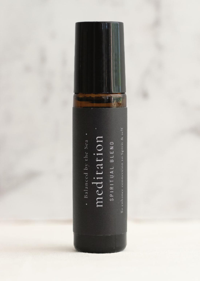 An amber essential oil roller bottle is displayed on a white surface with a white marble background. It has a black cap and a black Balanced by the Sea label with the words "Meditation - Spiritual Blend" on it