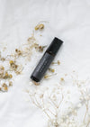 An amber essential oil roller bottle is lying on a white tablecloth, surrounded by chamomile and fine white gypsum flowers. It has a black cap and a black Balanced by the Sea label with the words "Meditation - Spiritual Blend" on it