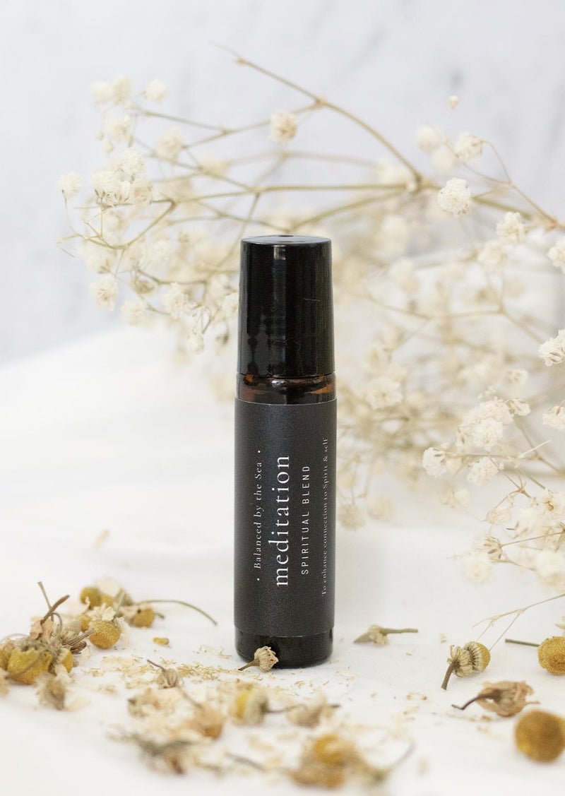 An amber essential oil roller bottle is standing on a white tablecloth, surrounded by chamolmile and fine white gypsum flowers. It has a black cap and a black Balanced by the Sea label with the words "Meditation - Spiritual Blend" on it