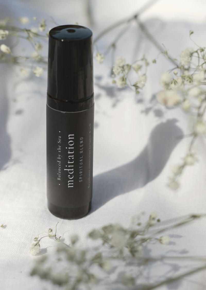 An amber essential oil roller bottle is standing on a white tablecloth, surrounded by fine white gypsum flowers. It has a black cap and a black Balanced by the Sea label with the words "Meditation - Spiritual Blend" on it