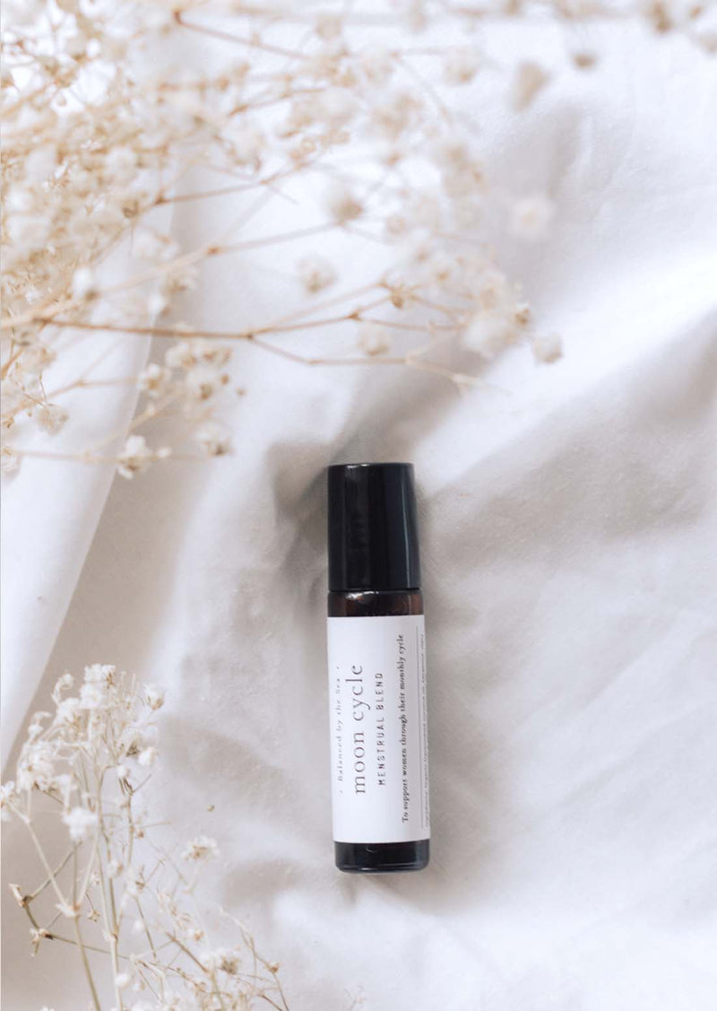 An amber essential oil roller bottle is lying on a white quilt, surrounded by fine white gypsum flowers. It has a black cap and a white Balanced by the Sea label with the words "Moon Cycle - Menstrual Blend" on it