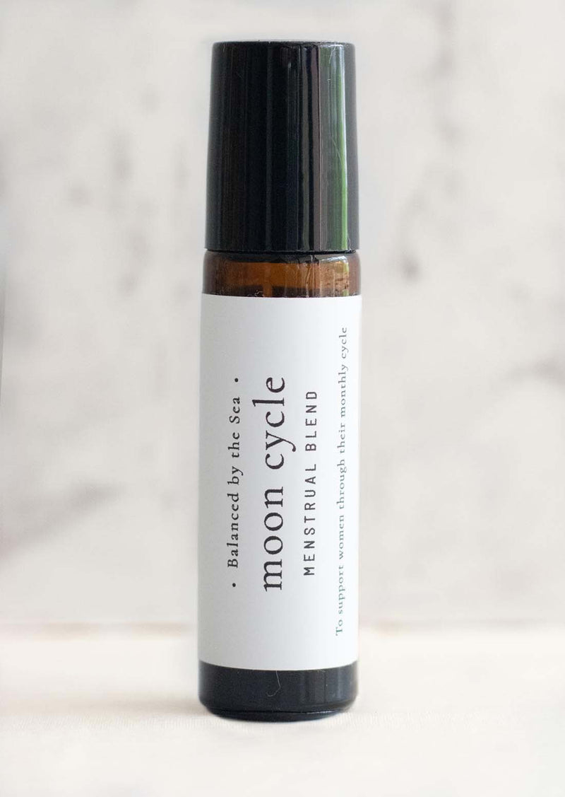 An amber essential oil roller bottle is displayed on a white surface with a white marble background. It has a black cap and a white Balanced by the Sea label with the words "Moon Cycle - Menstrual Blend" on it