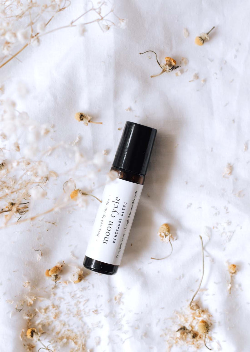 An amber essential oil roller bottle is lying on a white tablecloth, surrounded by chamomile and fine white gypsum flowers. It has a black cap and a white Balanced by the Sea label with the words "Moon Cycle - Menstrual Blend" on it