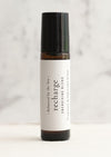 An amber essential oil roller bottle is displayed on a white surface with a white marble background. It has a black cap and a white Balanced by the Sea label with the words "Recharge - Energising Blend" on it