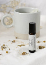 An amber essential oil roller bottle is lying on a white quilt, surrounded by chamomile flowers. It has a black cap and a white Balanced by the Sea label with the words "Recharge - Energising Blend" on it. There is a white ceramic coffee mug and some fine white gypsum flowers blurred in the background