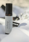 An amber essential oil roller bottle is standing on a white quilt. It has a black cap and a white Balanced by the Sea label with the words "Recharge - Energising Blend" on it. There are other roller bottles and some fine white gypsum flowers blurred in the background