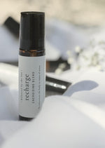 An amber essential oil roller bottle is standing on a white quilt. It has a black cap and a white Balanced by the Sea label with the words "Recharge - Energising Blend" on it. There are other roller bottles and some fine white gypsum flowers blurred in the background