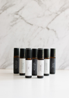 Nine amber Balanced by the Sea essential oil roller bottles are displayed on a white surface with a white marble background.