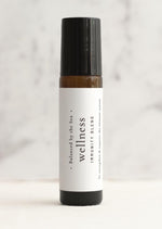 An amber essential oil roller bottle is displayed on a white surface with a white marble background. It has a black cap and a white Balanced by the Sea label with the words "Wellness - Immunity Blend" on it